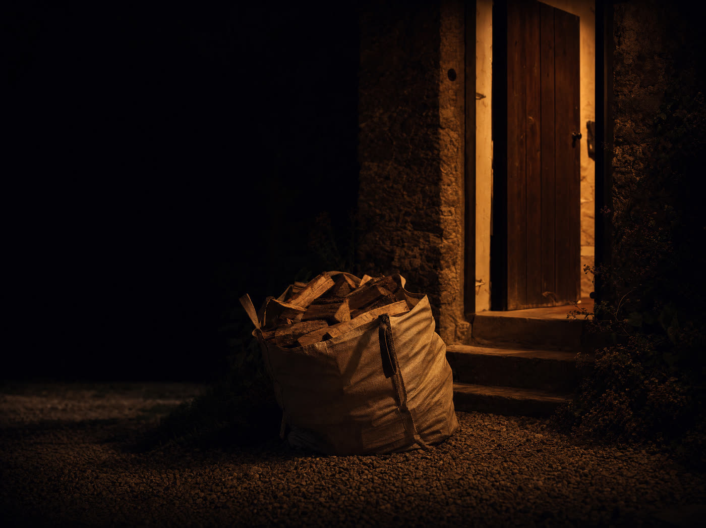 A woven bag of split logs set down on a gravel path at the foot of a front-door step